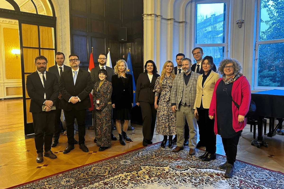 Grupa kilkunastu osób pozuje w reprezentacyjnej sali Ambasady RP w Wiedniu. W tle flagi Polski i Unii Europejskiej, fortepian oraz wysokie okna.